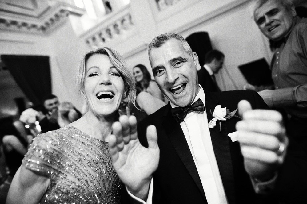 People smiling and celebrating at a formal event, dressed in tuxedo and gown, in a decorated indoor venue.