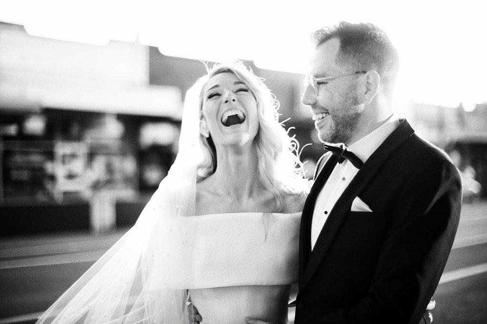 A bride and groom in wedding attire, smiling and laughing outside during daytime.