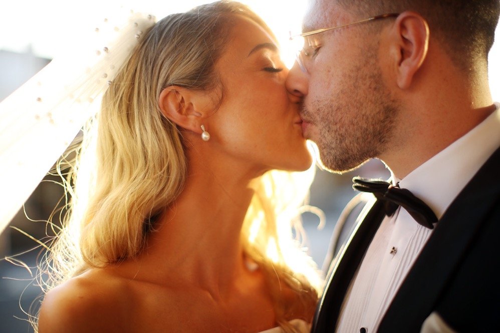 A bride and groom kiss, dressed in wedding attire, with sunlight in the background.