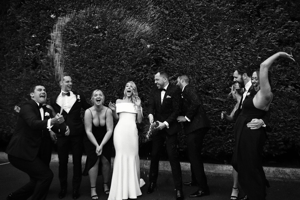 Group of people celebrating at a wedding, spraying and laughing, with a lush hedge in the background.