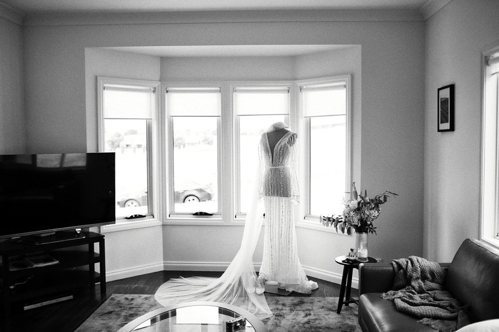 A bedroom with a wedding dress on a mannequin in front of a bay window, a TV on a stand, a sofa with a blanket, a small table with flowers, and framed wall art.