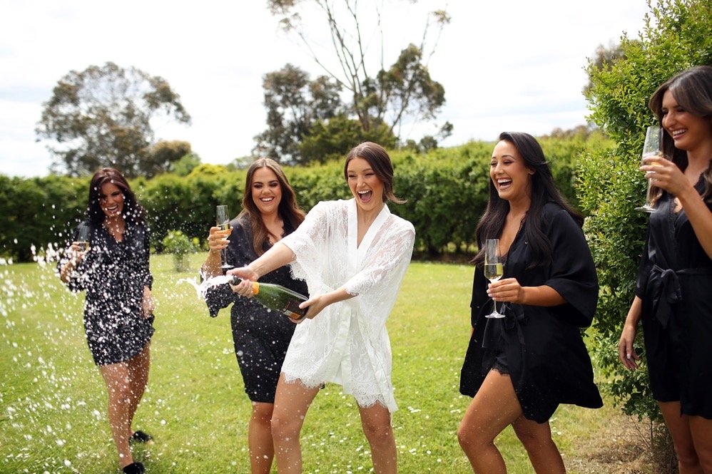 Group of women celebrating outdoors, with one opening a champagne bottle and others holding glasses, smiling and laughing.