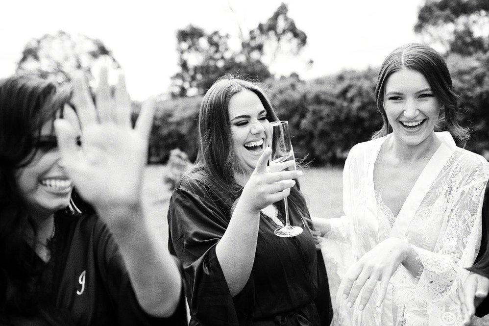 Three women outdoors, smiling and laughing, with one holding a glass of wine.