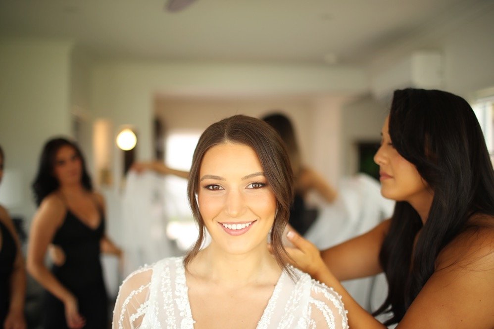 A young woman with brown hair and a white lace dress smiling as she gets her makeup and hair done by two women, with other women in black dresses in the background.
