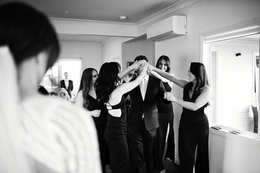 A group of women and a man dressed in formal attire celebrating indoors, with some women playfully covering the man's face.
