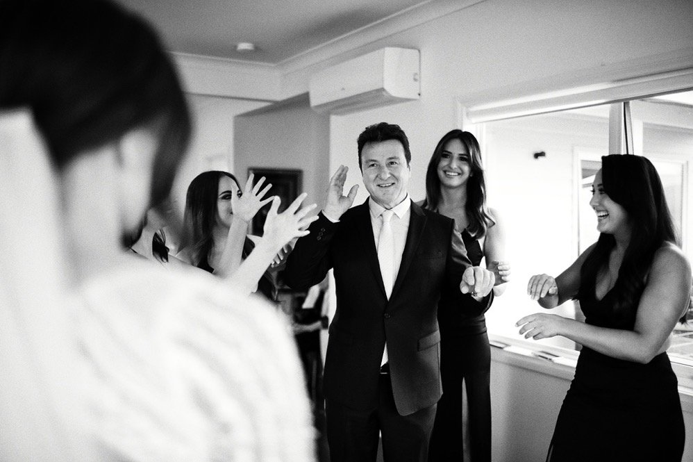 Group of people in formal attire enjoying a celebration indoors, with one man in a suit smiling and raising his hand.