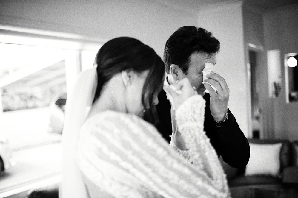 A bride and groom are emotional, holding tissues on their faces, possibly crying, inside a room.