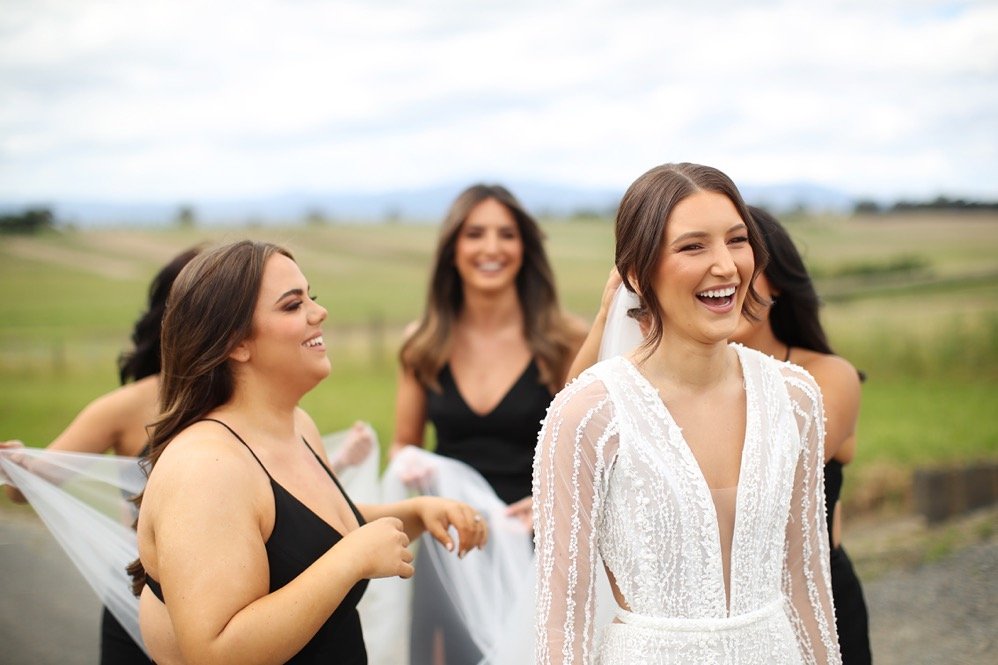 A bride in a white lace wedding dress smiling with her friends in black dresses outdoors on a cloudy day, countryside background.