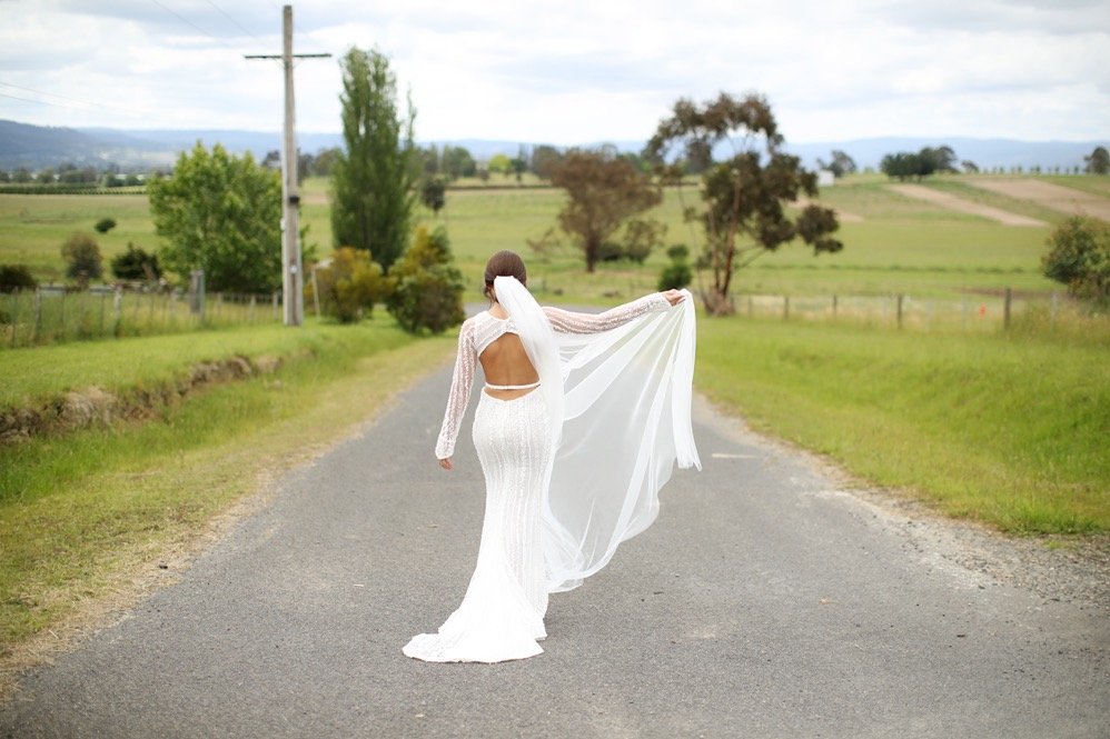 A bride in a white wedding dress standing on a country road, holding her veil as she faces away from the camera with a rural landscape in the background.