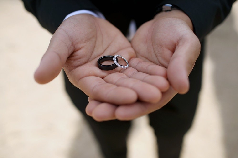 A person in a black suit holding two rings, one black band and one silver ring with diamonds.
