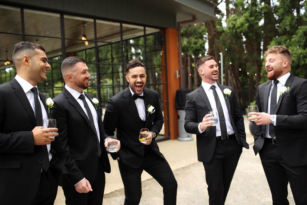Group of men in tuxedos and boutonnières, smiling and laughing while holding drinks at an outdoor wedding reception.