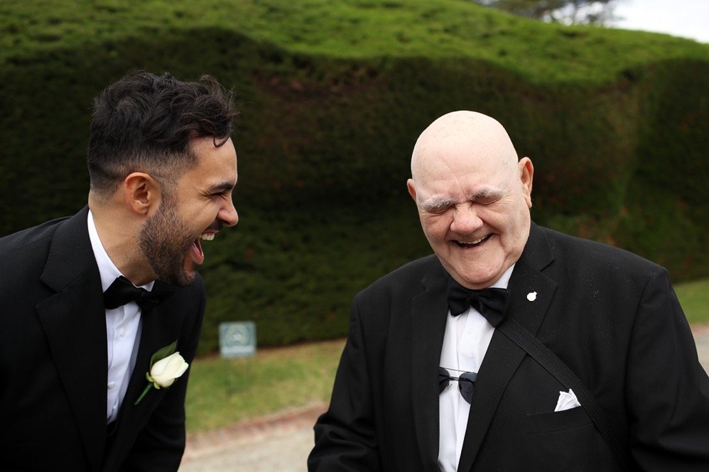 Two men in tuxedos laughing and smiling outdoors, with a grassy hill in the background.