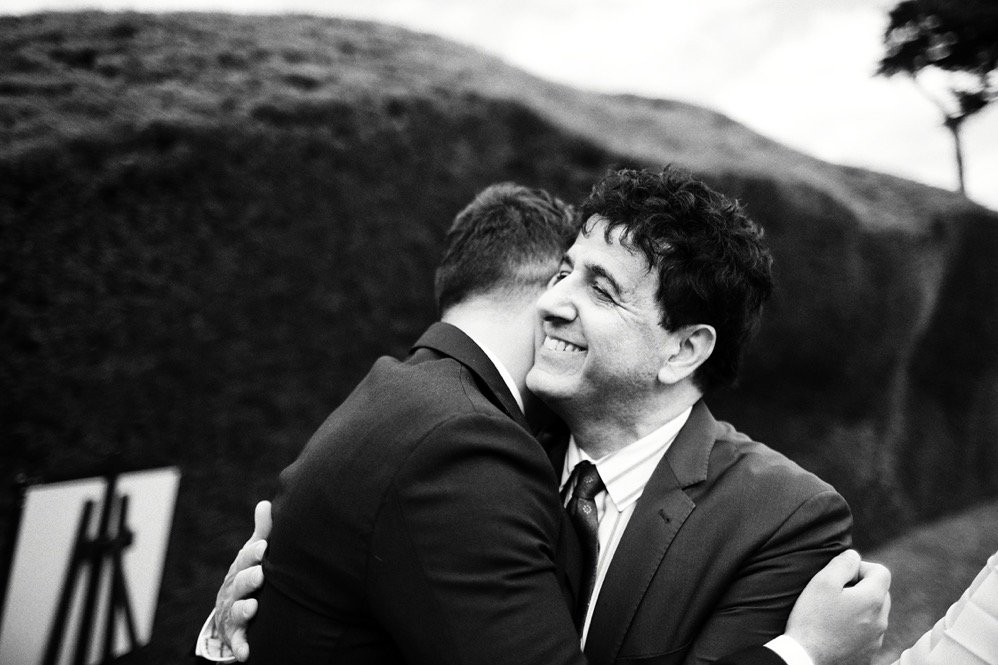 Two men in suits hugging outdoors, smiling, with a hill and trees in the background
