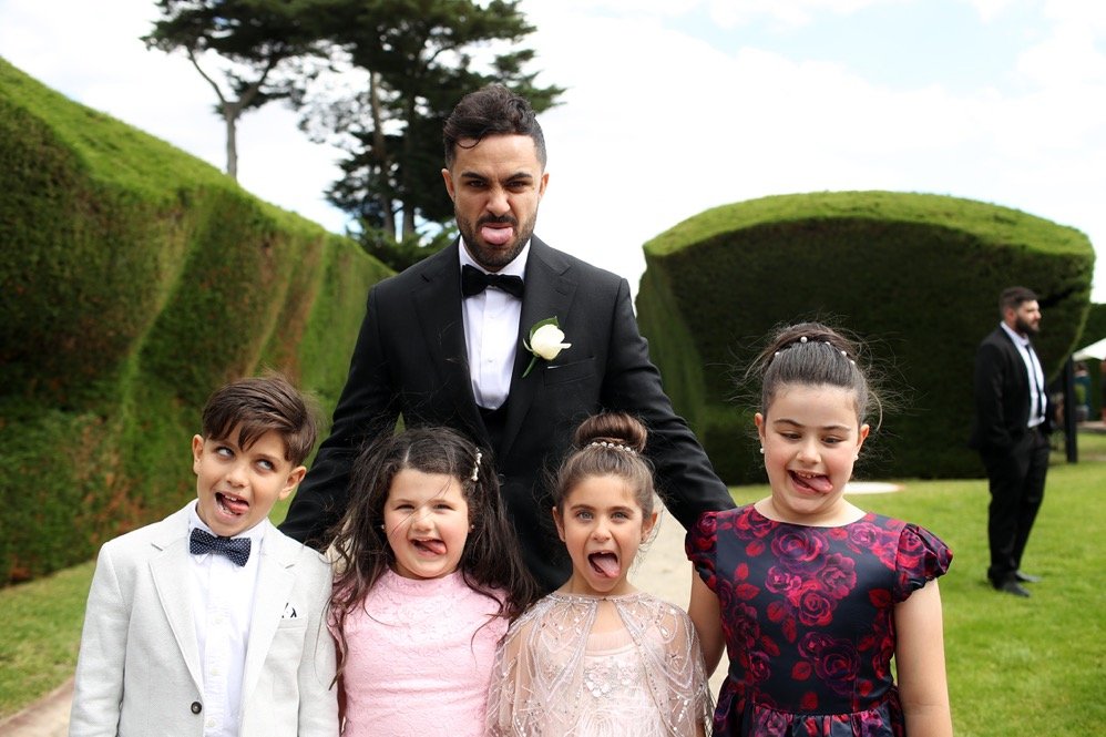 Group of children and a man making silly faces outdoors during a formal event, possibly a wedding, with greenery and trees in the background.