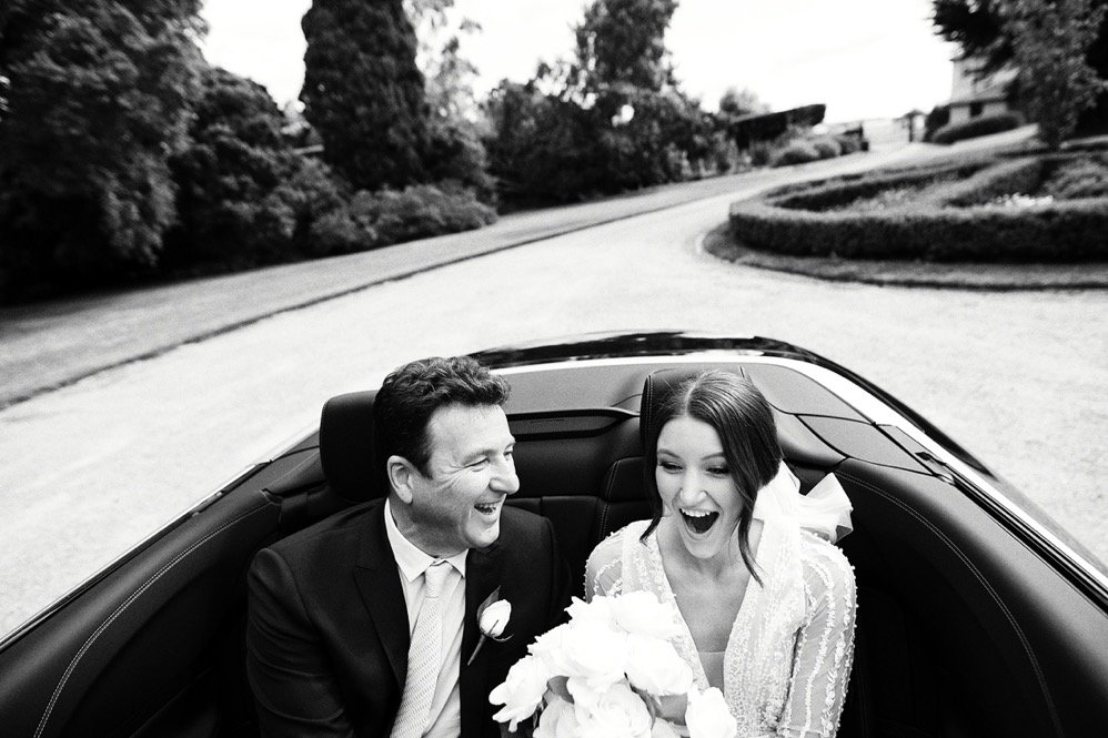 Black and white photo of a bride and groom sitting in the back of a convertible car on a driveway, smiling and laughing, with trees and bushes in the background.