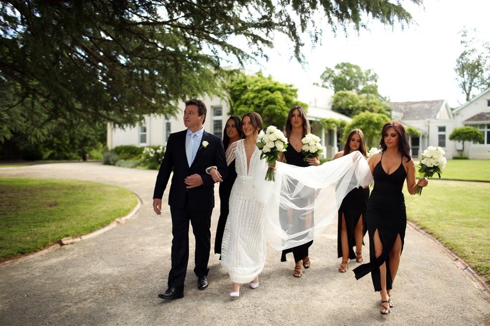 A bride in a white wedding gown and her escort, a man in a black suit, walking outdoors with four women in black dresses holding bouquets of white flowers. The women are walking behind the bride, and she is holding a small bouquet. They are on a curv