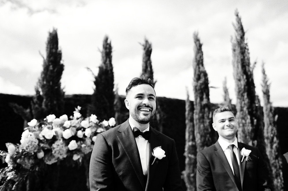 Two men in tuxedos with boutonnières, smiling at a wedding ceremony outdoors with cypress trees and flowers in the background.