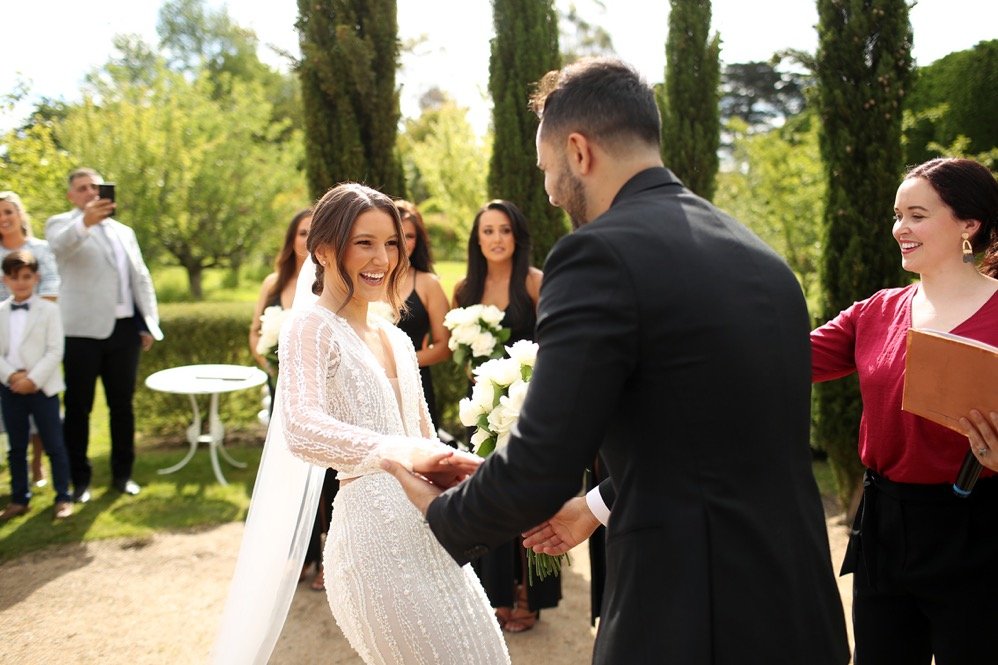 Bride and groom holding hands and smiling during a wedding ceremony outdoors, with guests watching and taking photos in the background.