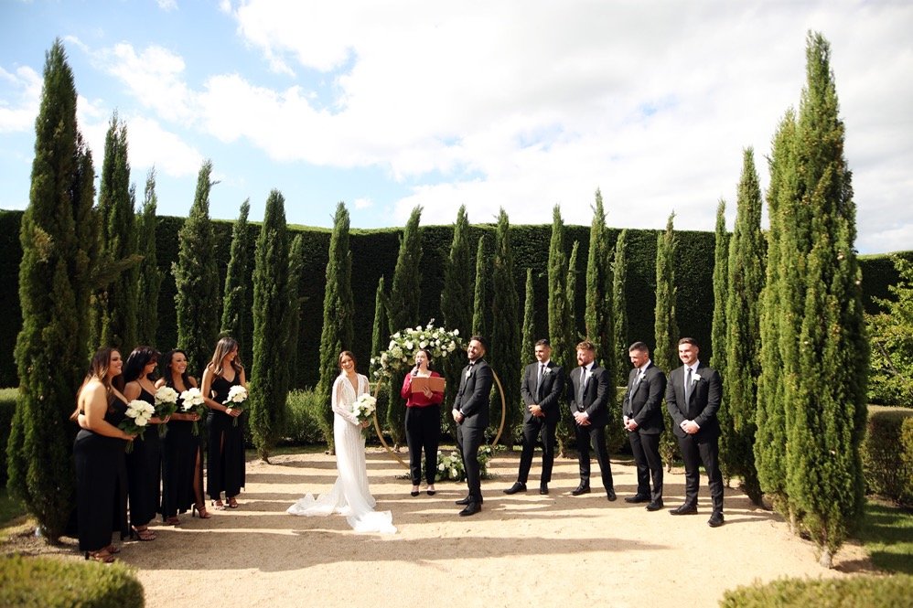 A wedding ceremony taking place outdoors in a garden with tall cypress trees, where the bride and groom are standing in front of a floral arch, surrounded by bridesmaids and groomsmen.