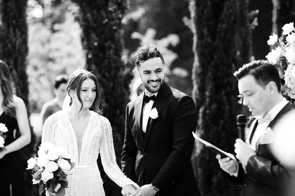 Black and white photo of a wedding ceremony outdoors, with a groom, bride, and officiant visible, surrounded by trees and flowers.