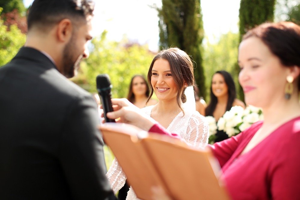 A wedding ceremony outdoors with a woman smiling at the officiant while holding a microphone, and another woman reading from a book. Bridesmaids are visible in the background.