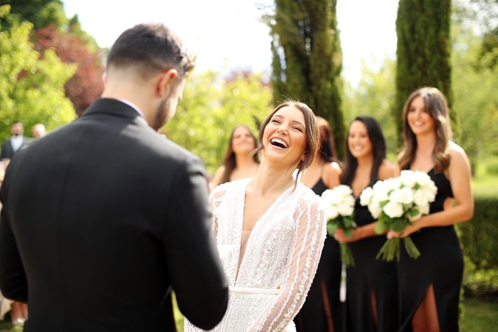 A bride and groom at an outdoor wedding ceremony, with bridesmaids holding bouquets in the background.