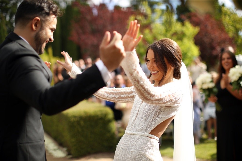 A bride and groom smiling and high-fiving outdoors during a wedding ceremony with guests in the background.