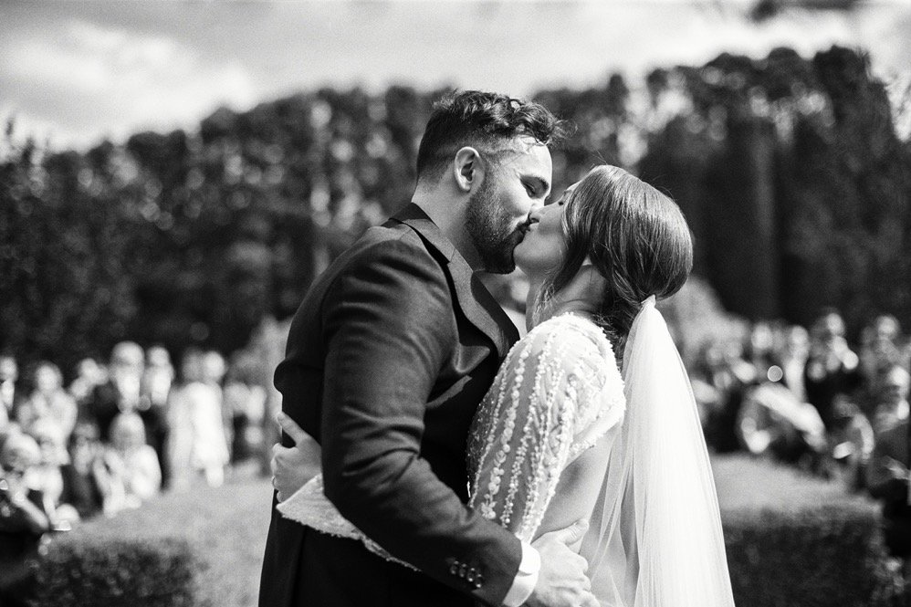 A couple kissing at their outdoor wedding, with guests in the background.