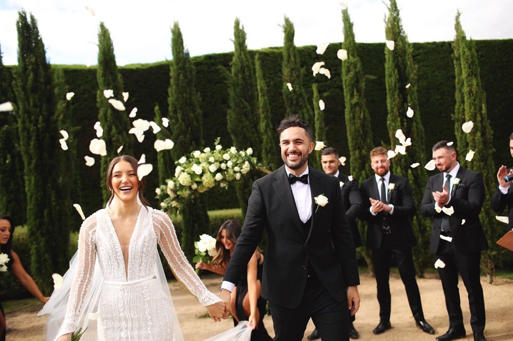 A newlywed couple walking hand in hand, smiling, with friends in suits and dresses behind them, releasing flower petals at an outdoor wedding in a garden with tall cypress trees.
