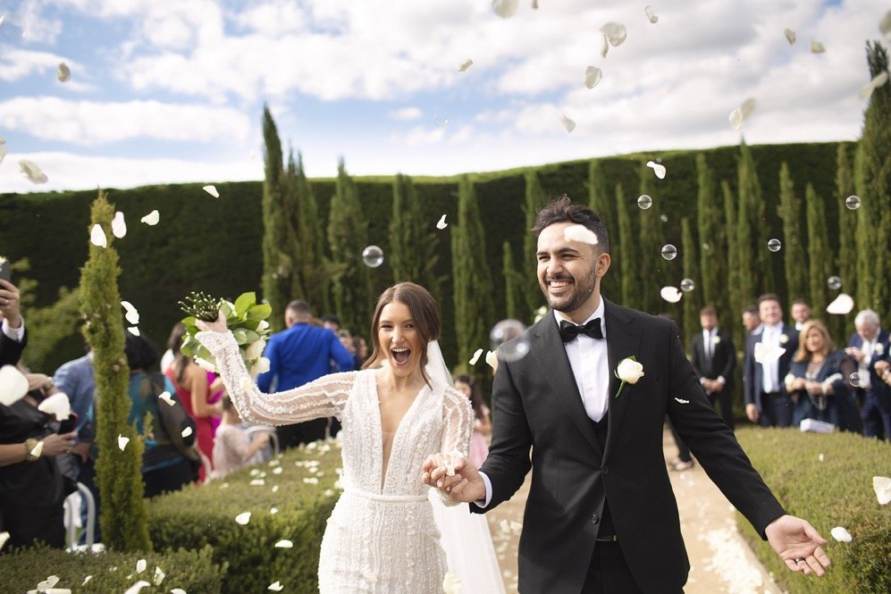 A bride and groom celebrating their wedding day outdoors with guests, surrounded by greenery, as rose petals and bubbles fill the air.