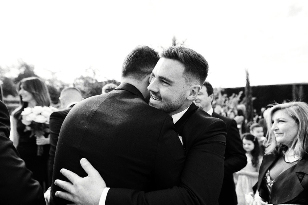 Two men in tuxedos embracing at a wedding or formal event outdoors, surrounded by other guests including women holding flowers and smiling.