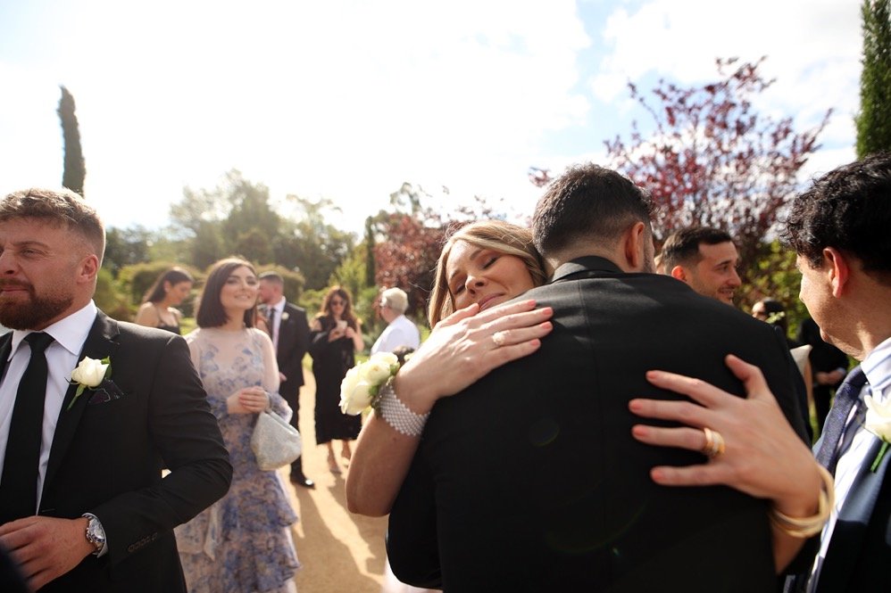 People at a wedding reception outdoors, with couples hugging and others smiling in the background.