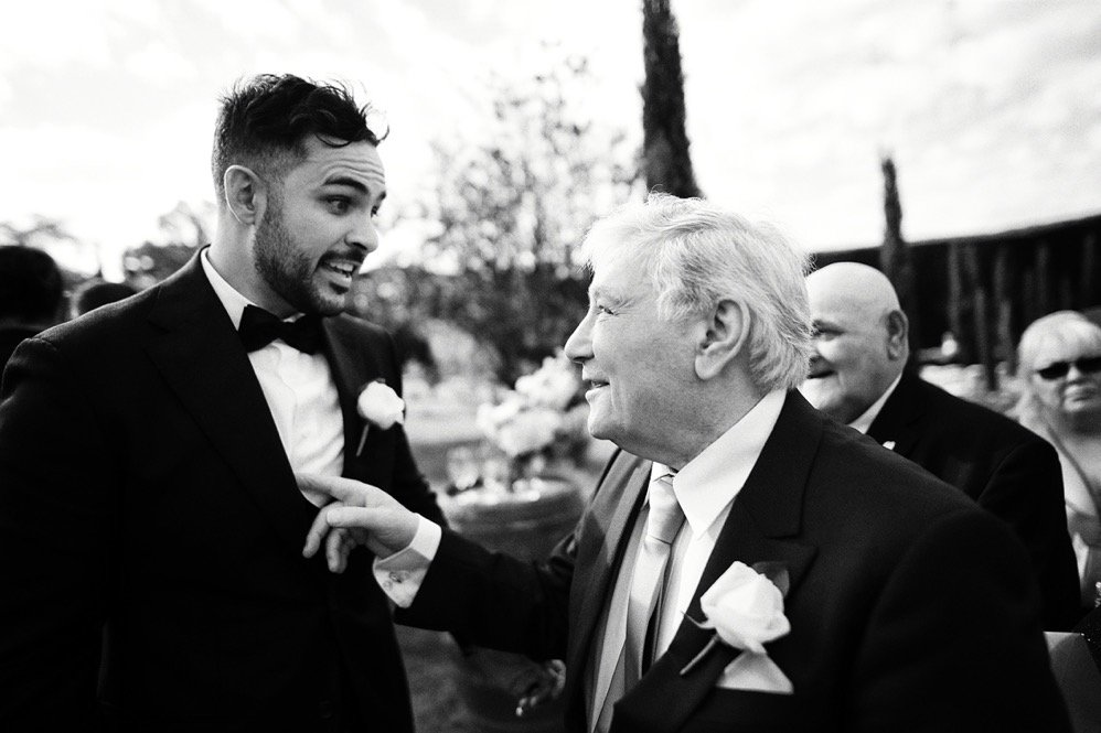 A young man in a tuxedo and an older man in a suit are happily talking and smiling at an outdoor social event, possibly a wedding, with other people in the background.