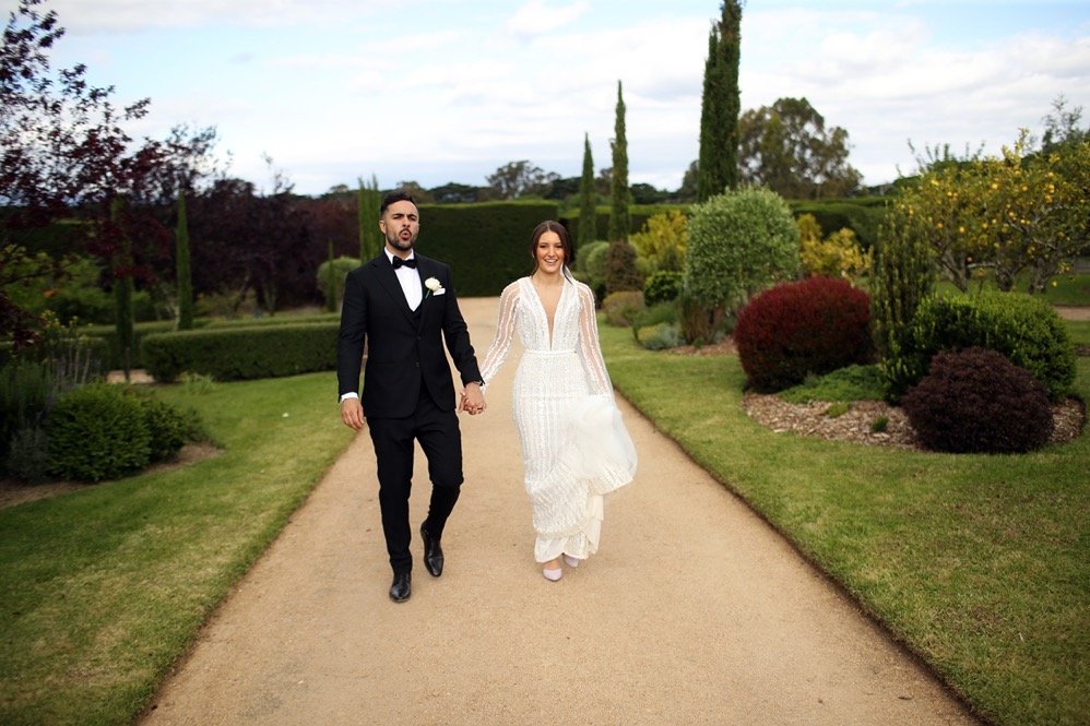 A bride and groom walking hand in hand on a garden path, smiling, with lush greenery and trees in the background, during a wedding photo shoot.