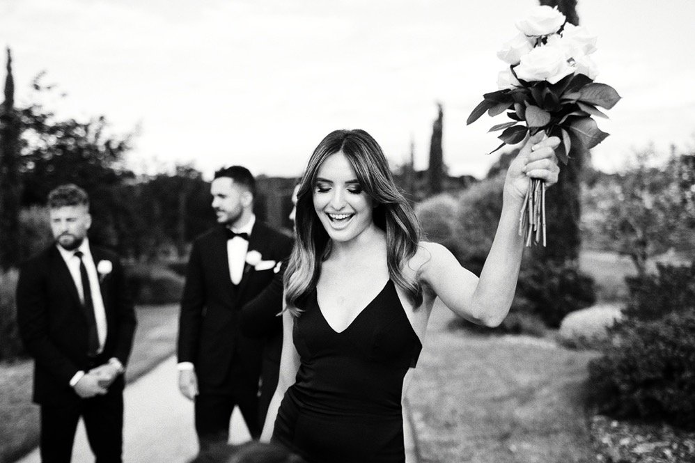 A woman in a black dress holding a bouquet of flowers, smiling, with three men in suits in the background outdoors.