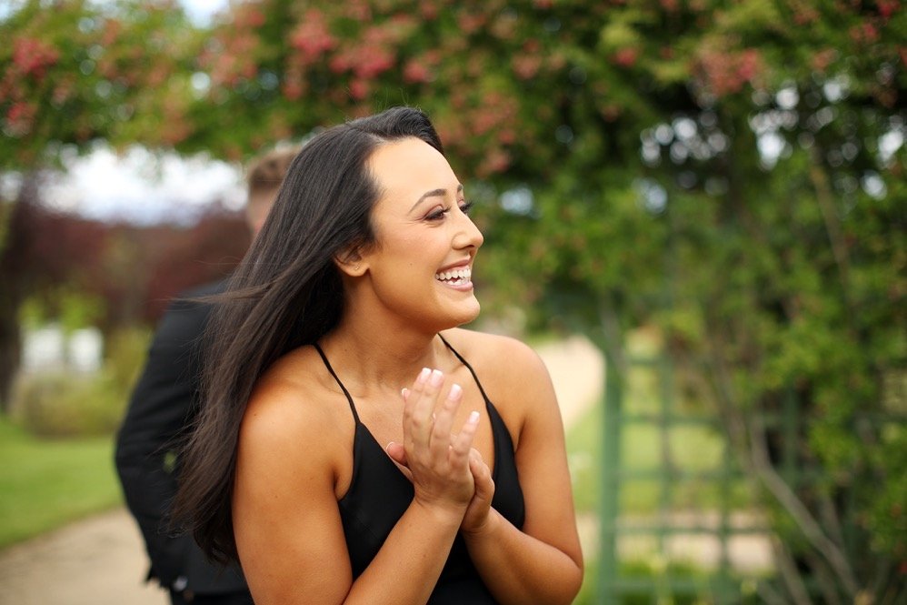 A woman with long dark hair wearing a black spaghetti strap dress, smiling and clapping outdoors with trees in the background.