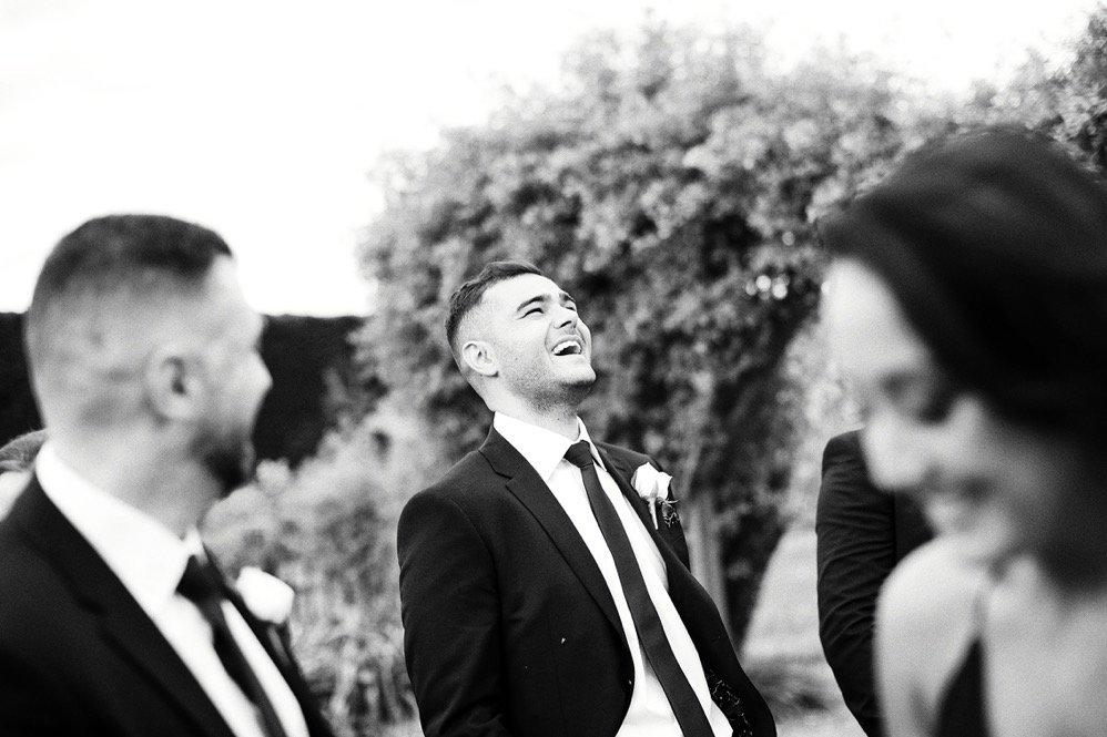 A group of people at a wedding outdoors, with the groom smiling and looking up, surrounded by others dressed in suits.