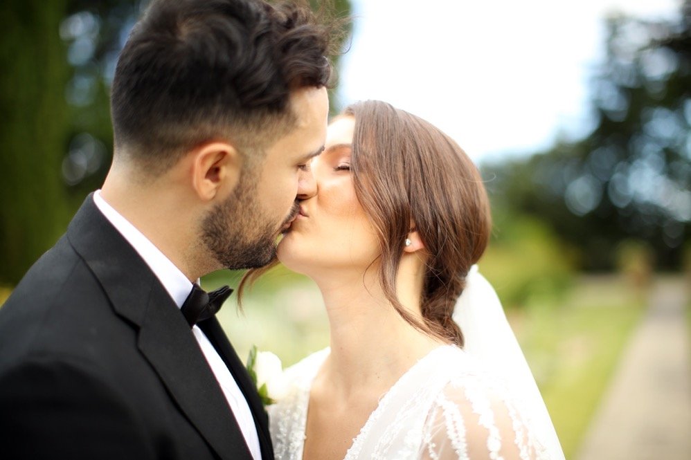 A newlywed couple sharing a kiss outdoors, with the bride in a white dress and the groom in a black tuxedo.