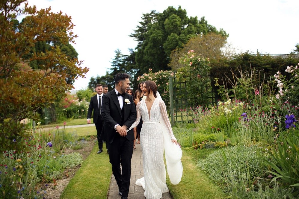 Bride and groom walking hand in hand through a garden, surrounded by colorful flowers, with wedding party in the background.