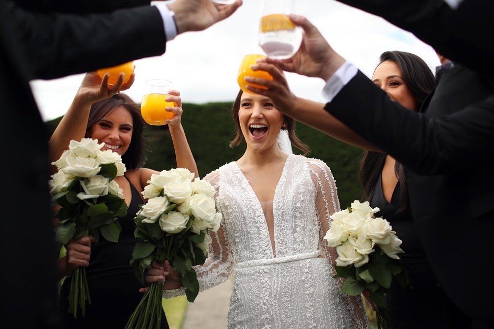 Group of women celebrating outdoors, holding bouquets of white roses and glasses of orange juice, with a bride in a white wedding dress smiling.