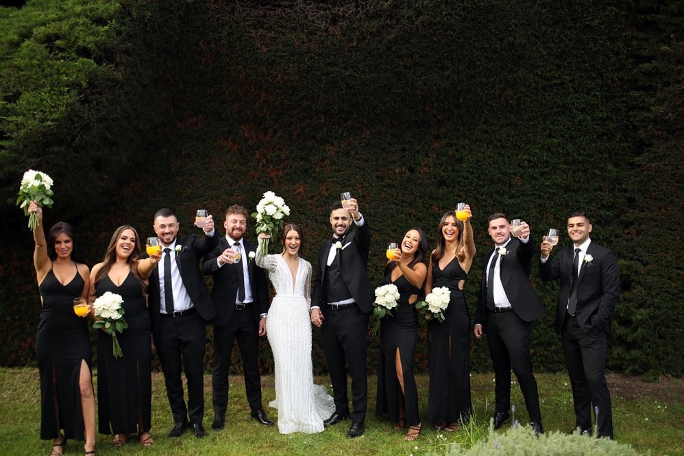Group of wedding guests and the bride and groom celebrating outdoors, holding flowers, drinks, and raising glasses in a toast.