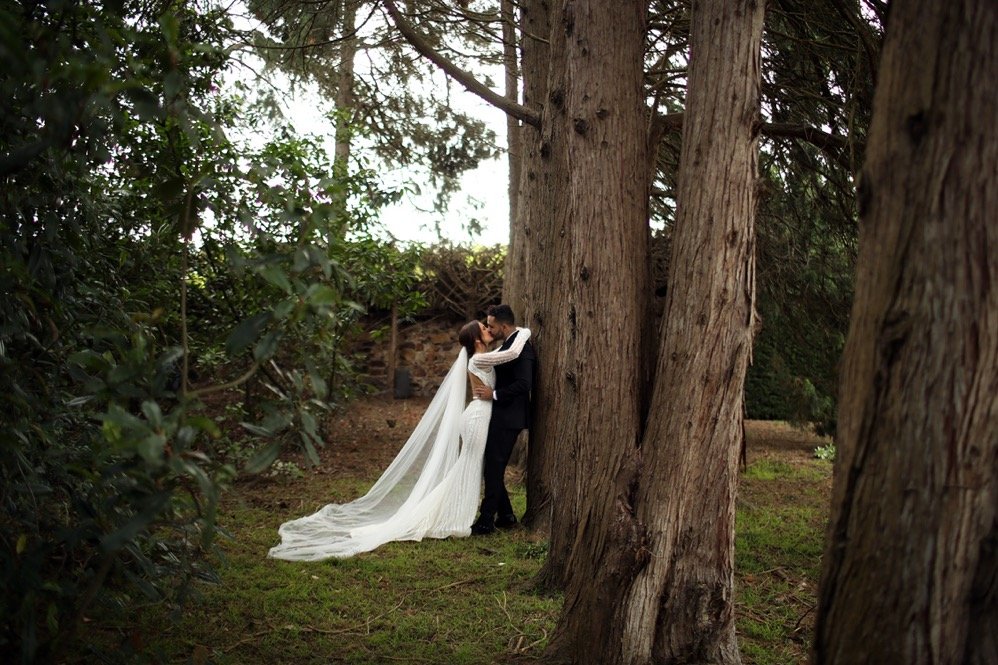 A bride and groom embrace in a forest, with tall trees and green foliage around them.