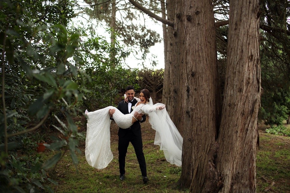 Groom lifting bride in a forested outdoor setting during a wedding photoshoot.