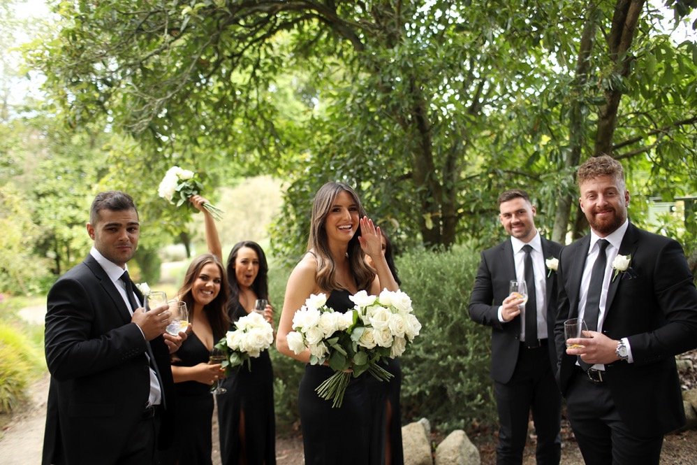 A group of six people dressed in formal attire, including three men and three women, gathered outdoors in a lush green park. They are holding glasses of beverages, and one woman is holding a bouquet of white flowers. They are smiling and appear to be