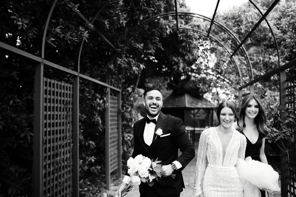 A joyful groom in a tuxedo holding a bouquet, walking with two smiling women in wedding dresses, outdoors behind a metal archway covered with trees.