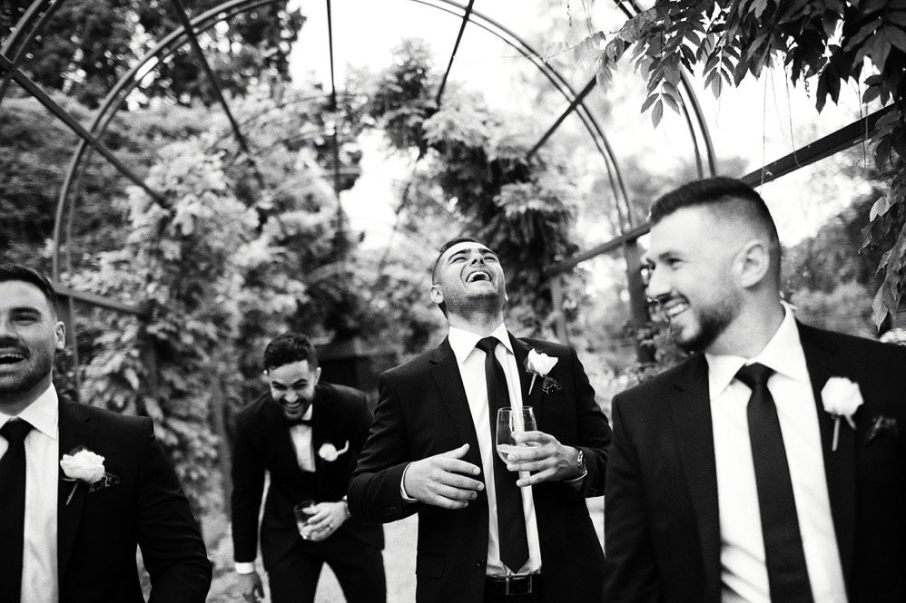 Group of men in suits laughing and smiling at a wedding or formal event in a garden or greenhouse setting.