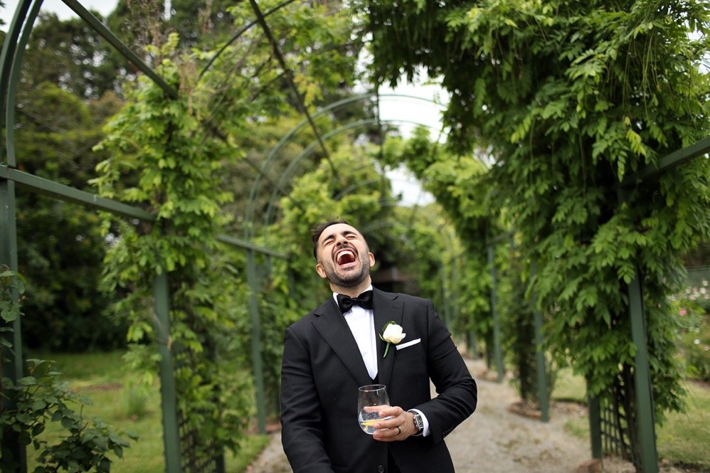Man in tuxedo laughing and holding a glass of drink in a lush green garden