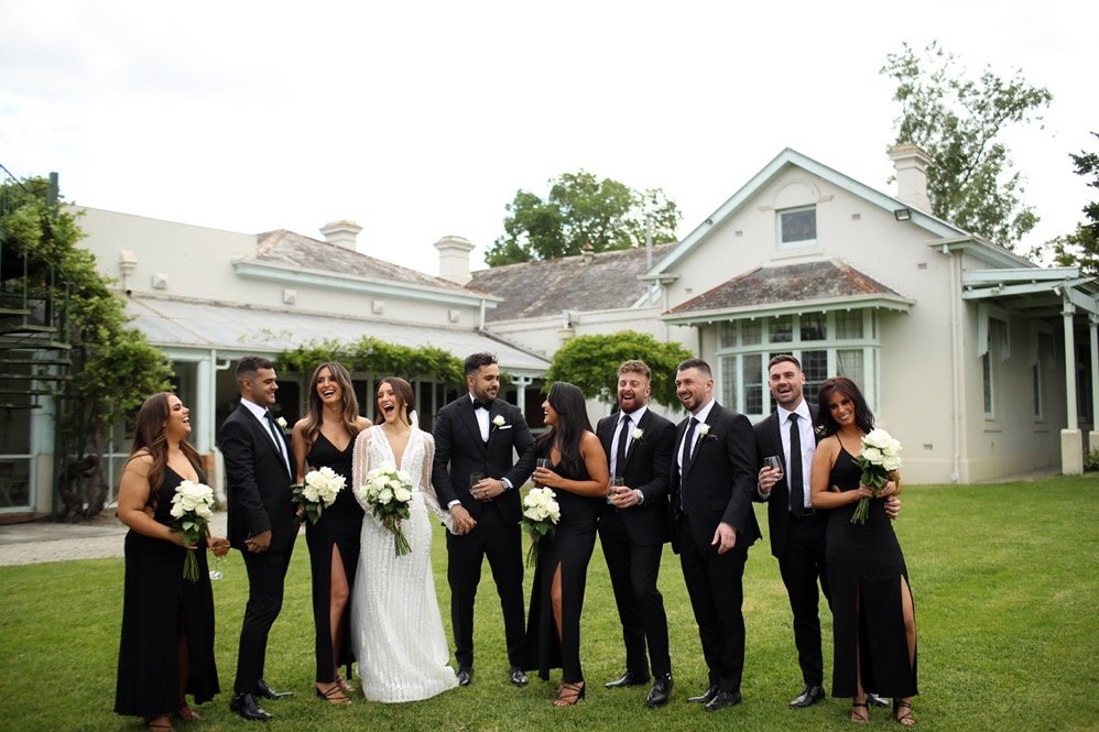 Group of wedding guests, including bridesmaids and groomsmen, standing on a lawn outside a house, celebrating and smiling, some holding flowers and drinks.