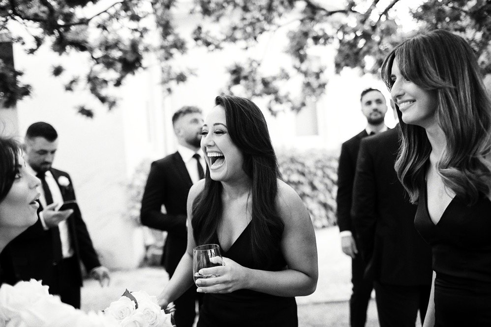 Black and white photo of women and men in formal attire enjoying a social gathering outdoors, with trees and a building in the background.