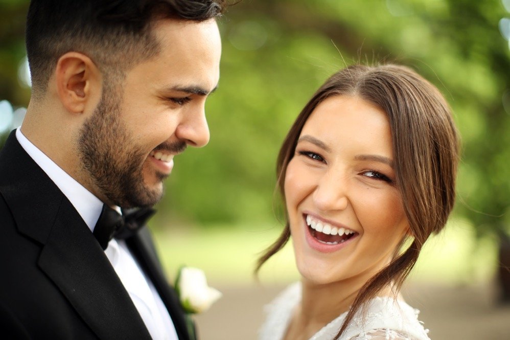 A smiling bride and groom in wedding attire outdoors, with trees and greenery in the background.